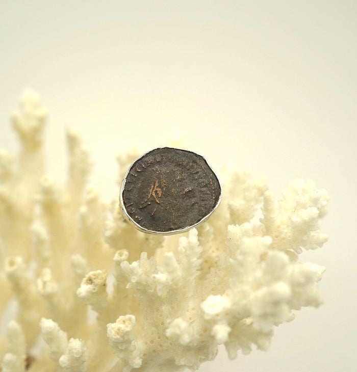 An antique bronze Roman coin set in a silver bezel, mounted on a silver band, displayed on a white floral-like surface.